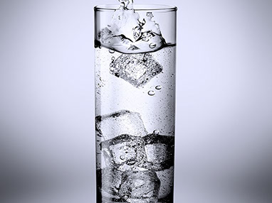 Un bicchiere d'acqua frizzante con il ghiaccio. Un spruzzo d'acqua provocato dal cubetto appena caduto dentro. Sfondo grigio freddo. Verticale.