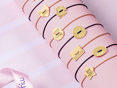 Braccialetti d'oro Mycharm su un tubo di cartoncino rosa. Nastro con il logo sulla sinistra in basso. Sfondo rosa con le strisce bianche.