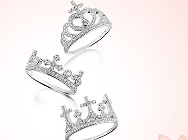 tre anelli d'argento con i cristalli, a forma di corona con le croci. Sfondo bianco-rosa e un fiore rosa in angolo basso destro. Verticale.