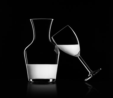 Una caraffa di vetro trasparente con il latte a sinistra e un bicchiere con il latte a destra, inclinato e appoggiato sulla caraffa. Sfondo nero