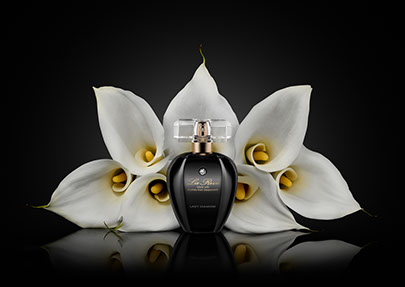 A black bottle of perfume in front of a symmetrical composition of white calla flowers. Black background.