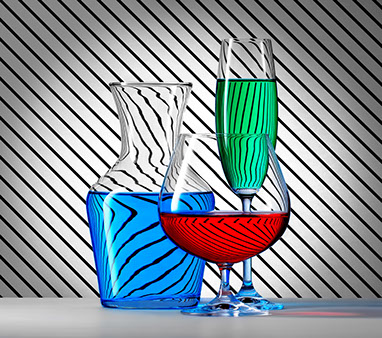 Una caraffa e due bicchieri di forme diverse, riempite con i liquidi colorati. Sfondo a strisce nere diagonali. Effetto di rifrazione.