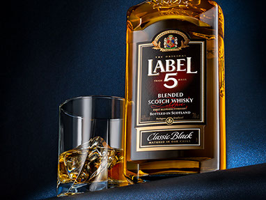 Bottiglia di whisky illuminata da dietro e  un bicchiere con il ghiaccio a sinistra. Sfondo blu perlato. Fotografato dal basso. Verticale