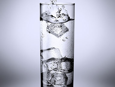 A glass of sparkling water with ice. A water splash caused by the cube just fell into it. Gray cold background. Vertical.