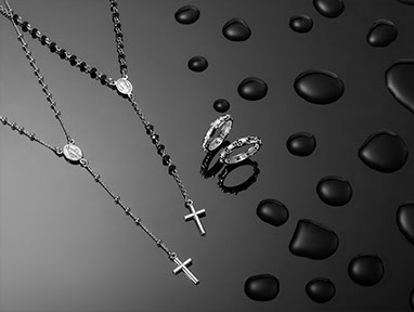 On the left two rosaries by Amen, on the right two rings. Silver and black crystals. Black base with drops of water on the right. Horizontal.