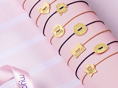 Gold bracelets by Mycharm on a pink cardboard tube. Ribbon with logo on the left bottom. Pink background with white stripes.