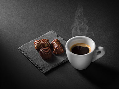 A cup of hot espresso, on the left three chocolates on a piece of black slate. Black background, the light coming from the top left.