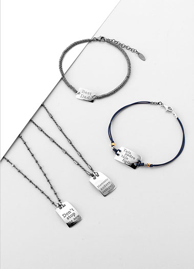 Due bracciali e due collane uomo in argento. I gioielli con le targhette con delle frasi. Sfondo bianco e grigio.