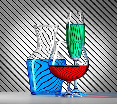 A carafe and two glasses of different shapes, filled with colored liquids. Black diagonal striped background. Effect of refraction. Horizontal.