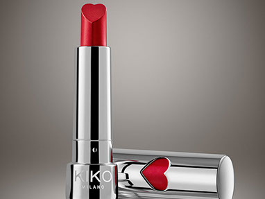 A red lipstick by Kiko, in the chrome package with a red heart. The stick is heart shaped. Beige background. Horizontal.