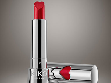 A red lipstick by Kiko, in the chrome package with a red heart. The stick is heart shaped. Beige background. Horizontal.