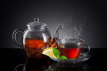 On the left a glass teapot with hot blooming tea, to the right a clear glass tea cup with lemon slice and mint leaf on the saucer.