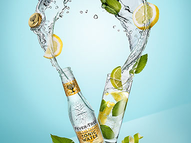 A dynamic composition with a bottle of tonic water and a glass, with liquid splashes, flying pieces of lemon and mint leaves